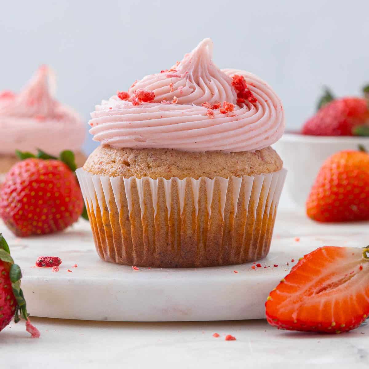 Close up of a strawberry cupcake with pink strawberry cream cheese frosting surrounded by fresh strawberries
