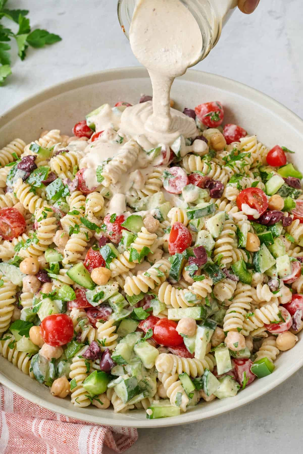 Tahini dressing being poured onto a big bowl of tahini pasta salad with tomatoes, cucumbers, bell pepper, chickpeas, and olives.