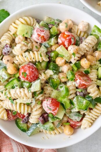 Closeup of a small bowl of tahini pasta salad with tomatoes, cucumbers, chickpeas, bell pepper, parsley, and olives.
