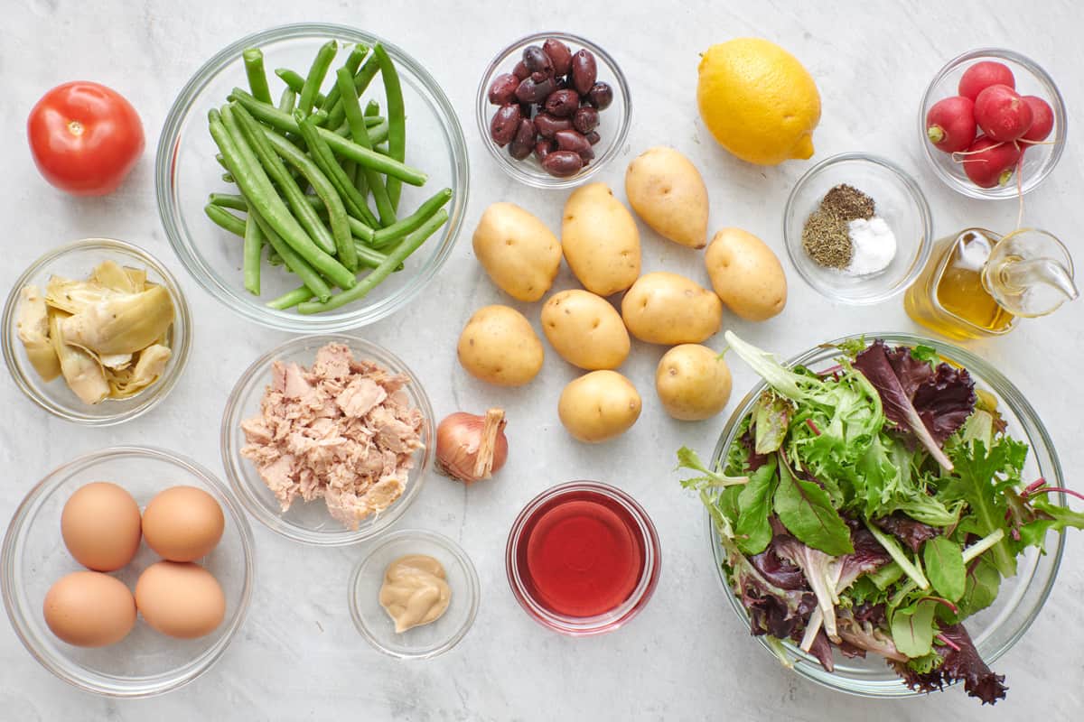 Ingredients for recipe before prepping and in individual bowls: tomato, artichoke hearts, eggs, fresh green beans, tuna, dijon mustard, olives, potatoes, onion, red wine vinegar, lemon, spices, spring mix lettuce, radishes, and oil.