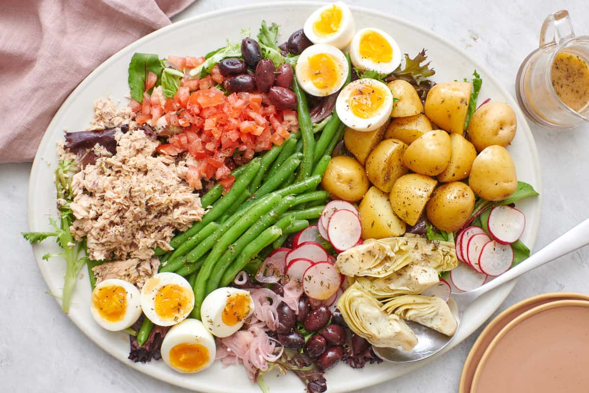 Tuna Nicoise Salad on a large platter with chopped tomatoes, green beans, hard boiled eggs, sliced radishes and shallot, quartered potatoes, artichoke hearts and lettuce with a side of dressing.