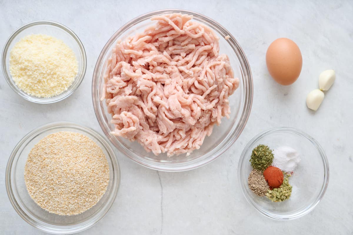 Ingredients for recipe in individual bowls before prepping: parmesan, breadcrumbs, ground chicken, egg, garlic, and spices.