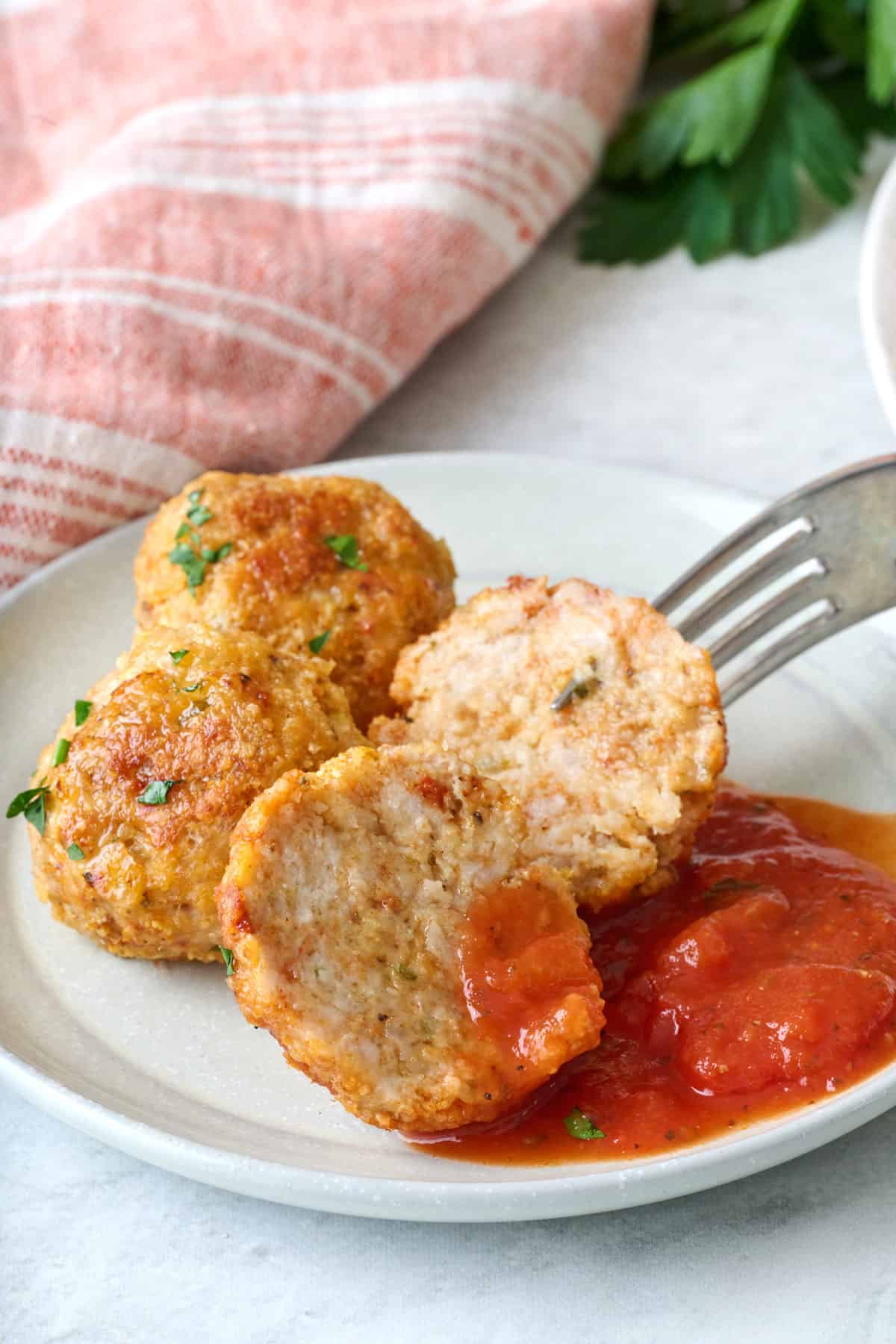 A few chicken meatballs with parmesan on a plate with one cut in half to show inside texture, marinara on plate with meatballs.
