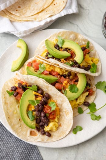 A plate of 3 breakfast tacos with eggs, salsa, beans, and avocado.