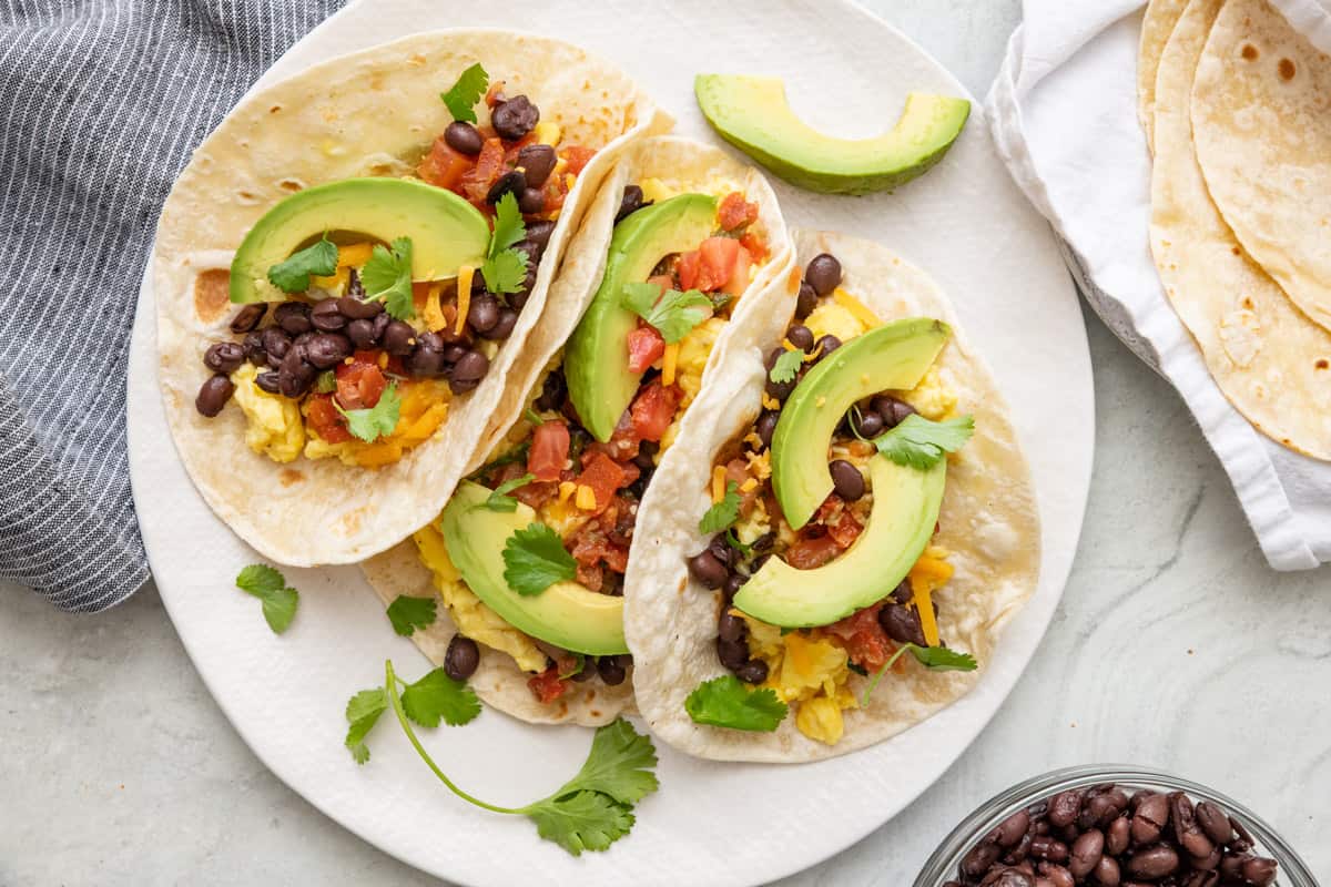 Overhead of breakfast tacos on a plate with eggs, cheese, salsa, beans, and avocado.