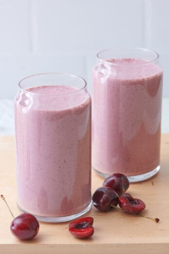 2 glasses of cherry smoothie on a table with cherries nearby.
