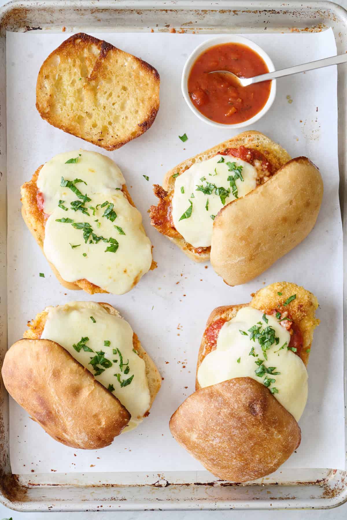 Chicken parm sandwiches on a parchment-lined baking sheet with top rolls half on and a small bowl of marinara with a spoon.