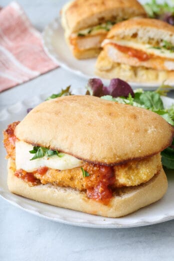 Closeup of a chicken parmesan sandwich on a small white plate with a sandwich cut in half in the background.