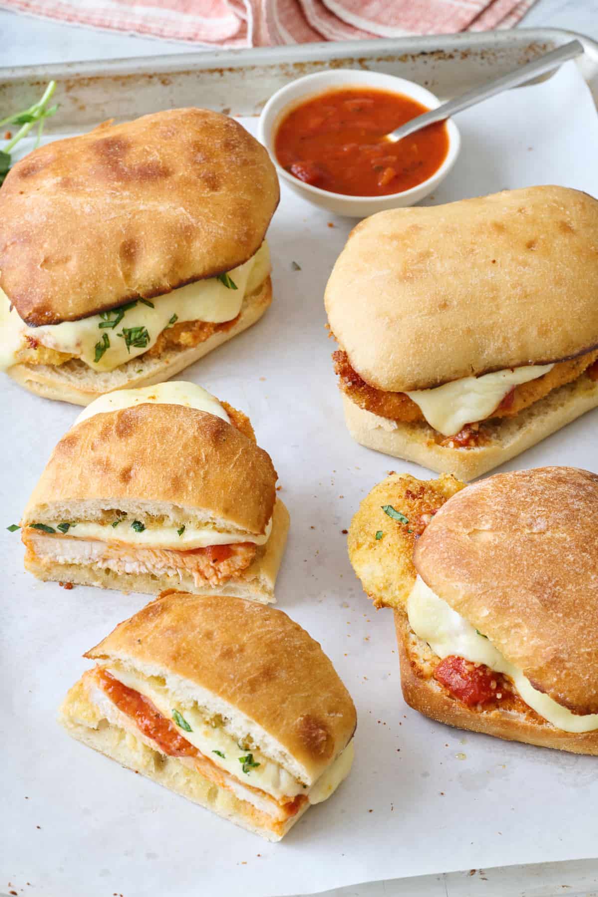 Chicken parm sandwiches on a parchment-lined baking sheet with a small bowl of marinara.