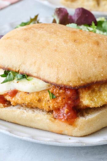 Closeup of a chicken parm sandwich on a small white plate with a side salad.