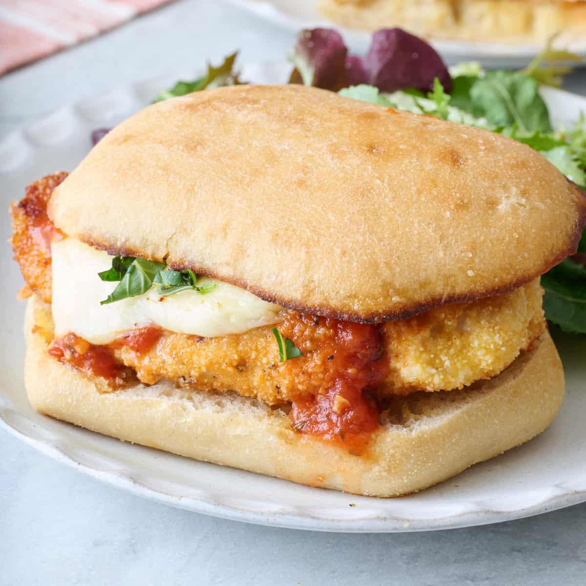 Closeup of a chicken parm sandwich on a small white plate with a side salad.