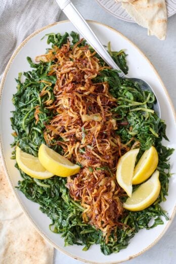 Dandelion greens recipe on a platter topped with crispy fried onions and lemon wedges with a spoon dipped into the side.