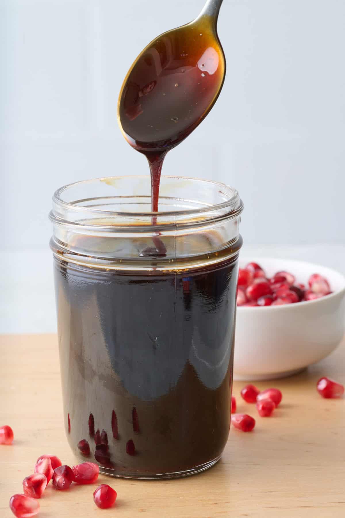 Spoon drizzling pomegranate molasses back into jar from above. Small bowl of pomegranate seeds nearby.