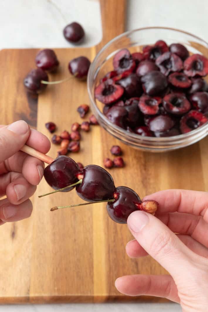 Hand holding a chopstick with 2 cherries on it and pushing the pit out of a 3rd cherry over a cutting board with pitted cherries in a bowl in the background.