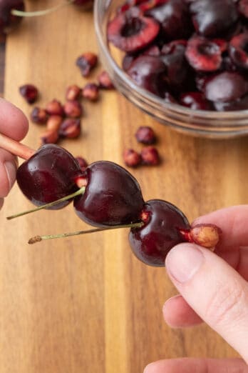 Hand holding a chopstick with 2 cherries on it and pushing the pit out of a 3rd cherry; cutting board with pitted cherries next to pits in the background.