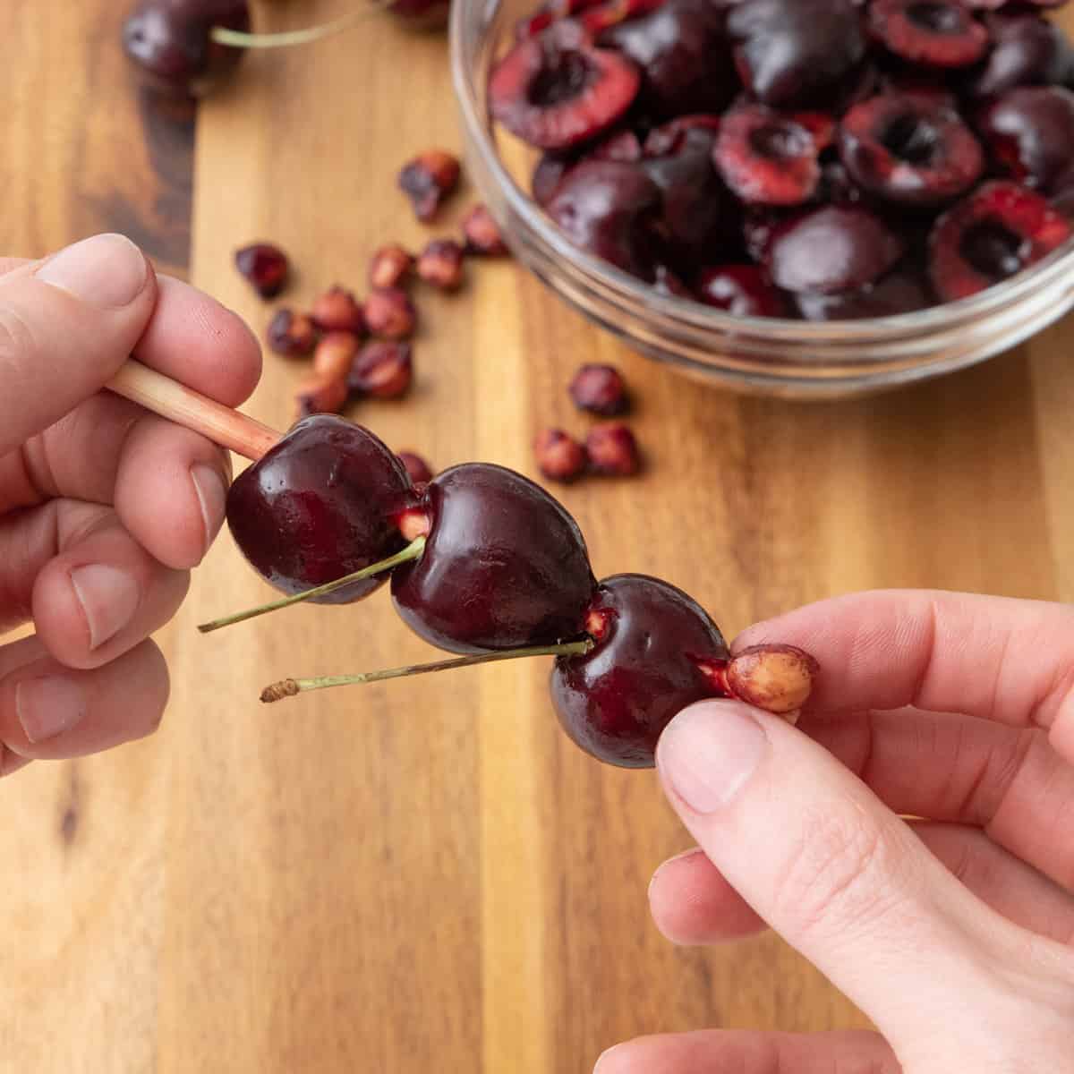 Hand holding a chopstick with 2 cherries on it and pushing the pit out of a 3rd cherry; cutting board with pitted cherries next to pits in the background.
