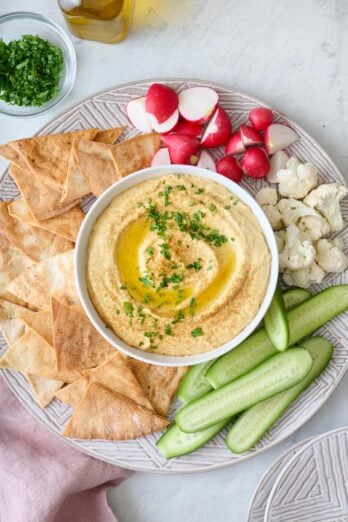 Hummus recipe without tahini in a small bowl drizzled with olive oil and sprinkled with cumin and fresh parsley served with pita chips and fresh veggies on a platter.