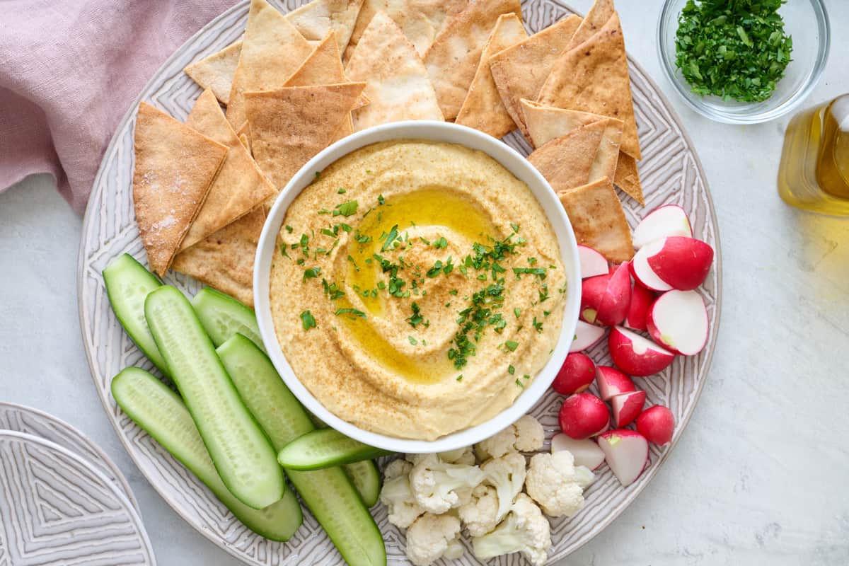 Hummus in a small white bowl with a drizzle of olive oil, sprinkle of cumin, and fresh parsley garnish served with fresh veggies and pita chips.