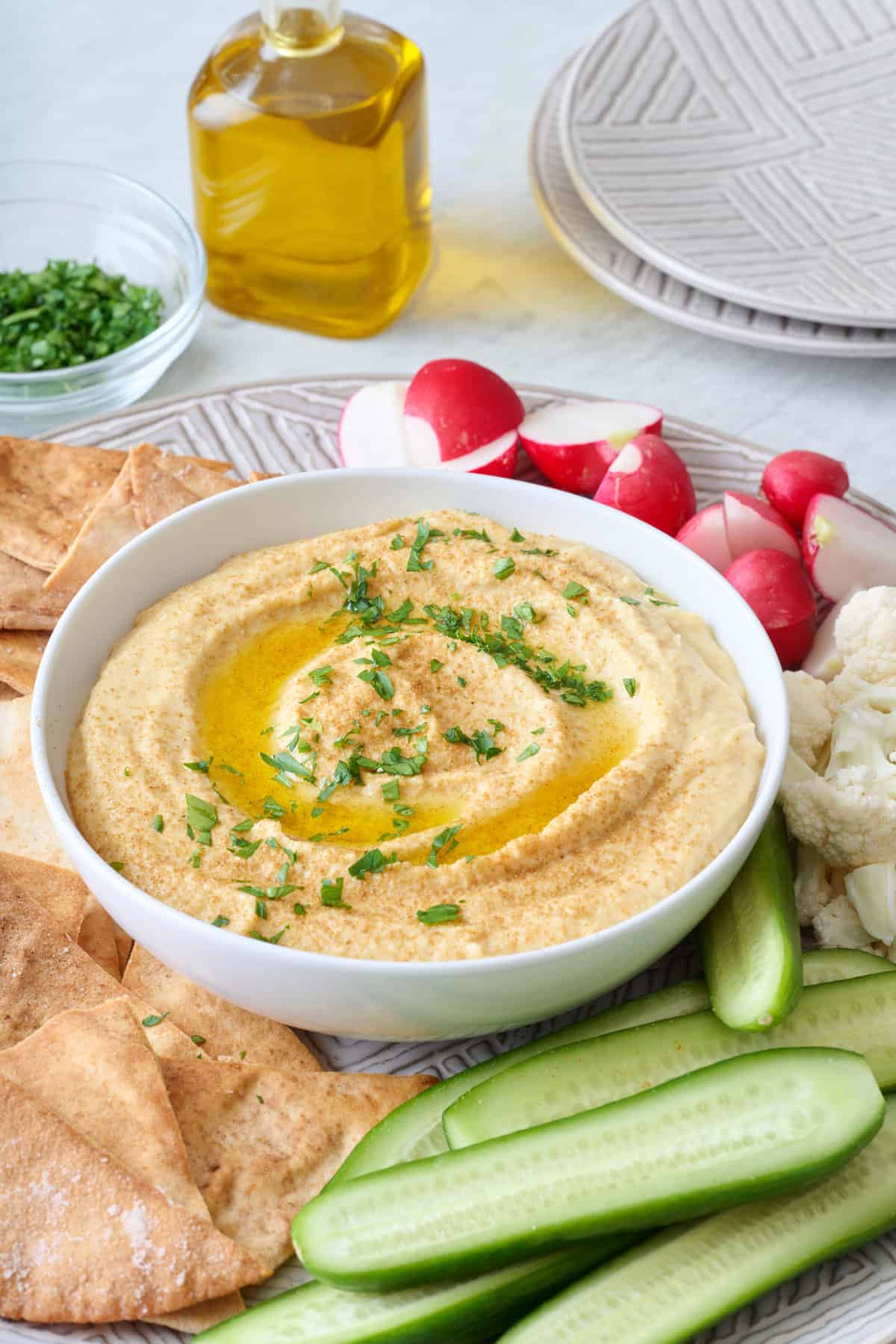 Hummus recipe without tahini in a small white bowl served on a platter with pita chips, cucumbers, radishes, and cauliflower.