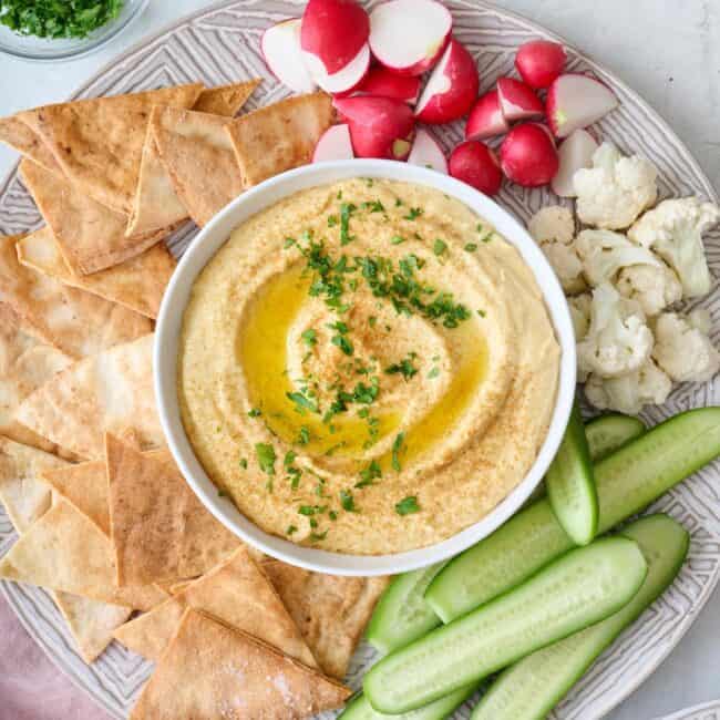 Homemade hummus recipe without tahini served with pita chips, cucumber, cauliflower, and radishes.