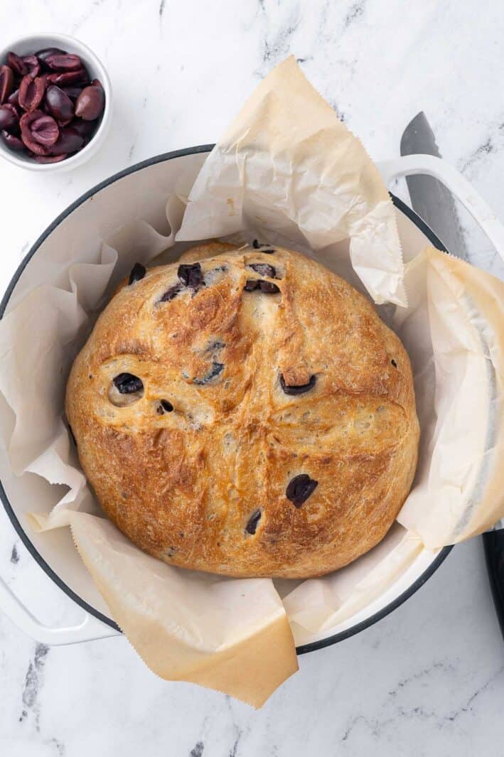 Baked olive bread in a parchment-lined dutch oven with a bread knife and small bowl of olives nearby.