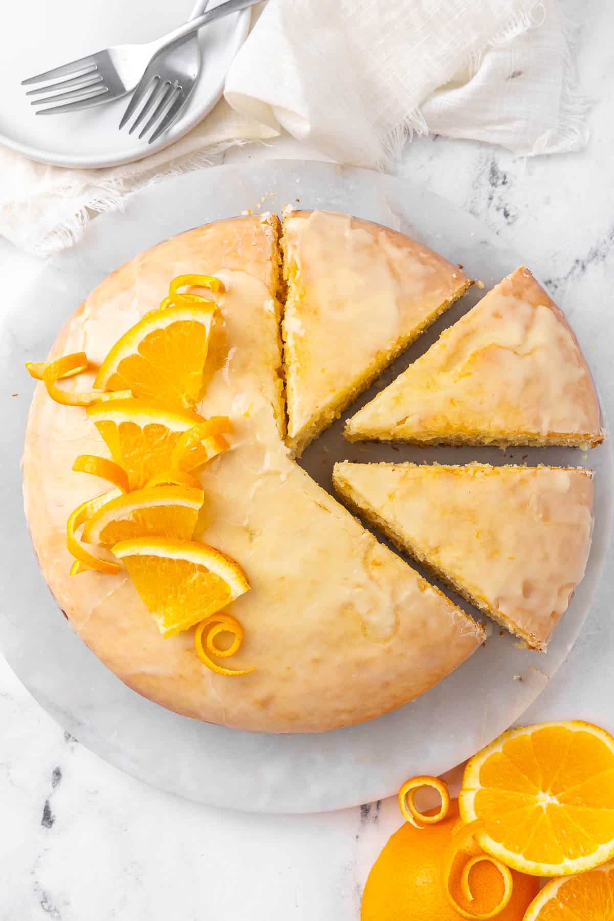 Orange cake on a marble plate with 3 slices cut out; napkin, forks, small plate, and orange slices nearby.