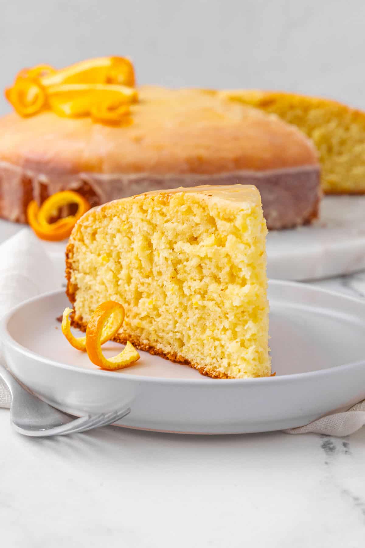 Closeup of a slice of orange cake on a white small plate with a fork and the whole cake in the background.