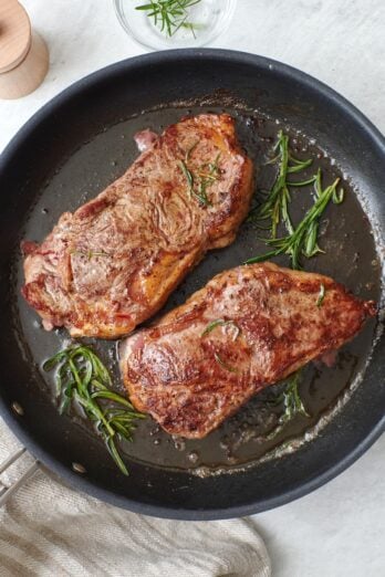 NY strip steaks on a non-stick skillet with melted butter spooned on top and fresh rosemary sprigs.