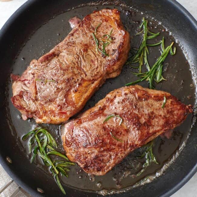 Closeup of pan-seared steaks in a skillet with sprigs of rosemary.