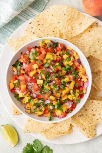 Overhead of a bowl of peach salsa surrounded by tortilla chips, fresh cilantro, a peach, and a lime slice.