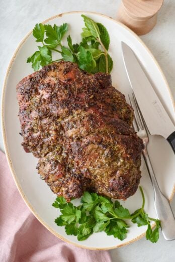 Roasted leg of lamb on a platter with fresh parsley, a fork, and a carving knife.