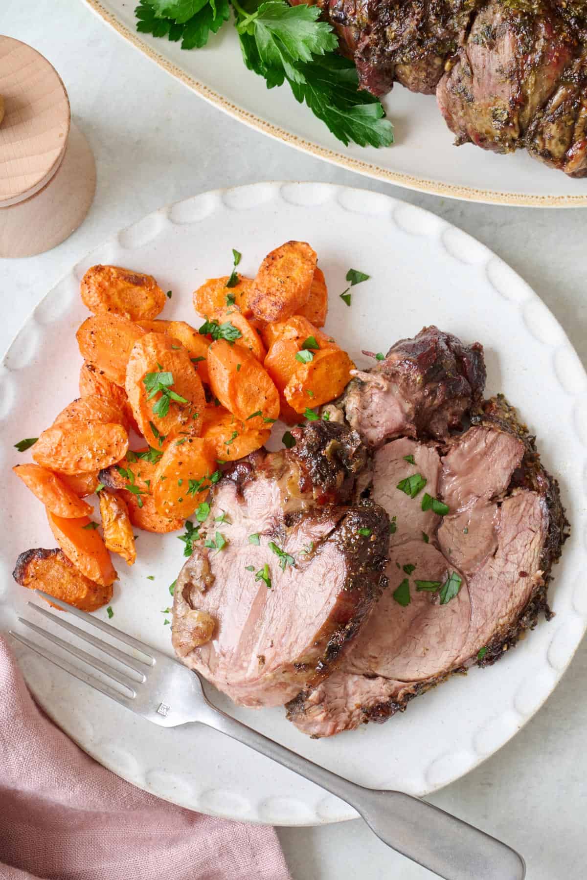 Slices of roasted leg of lamb on a white plate with roasted carrots and a fork.