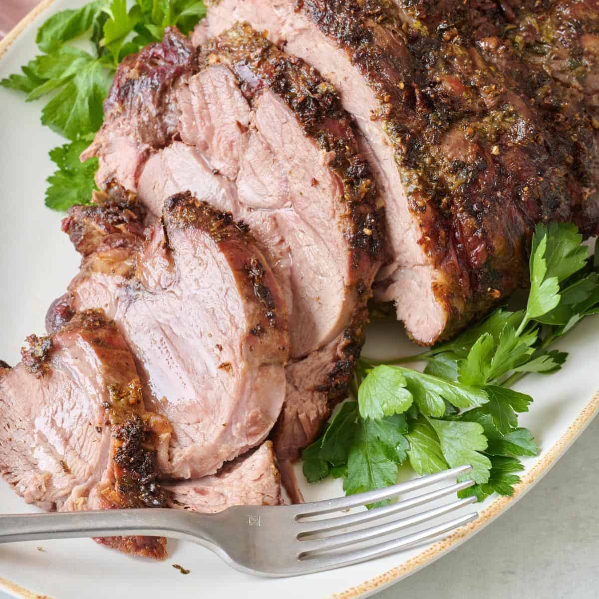 Sliced roasted leg of lamb on a plate with fresh parsley garnish and a fork.