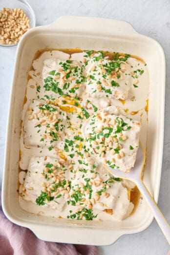 Samke harra with tahini sauce, toasted pine nuts, and chopped parsley in a baking dish with a spatula removing a portion.