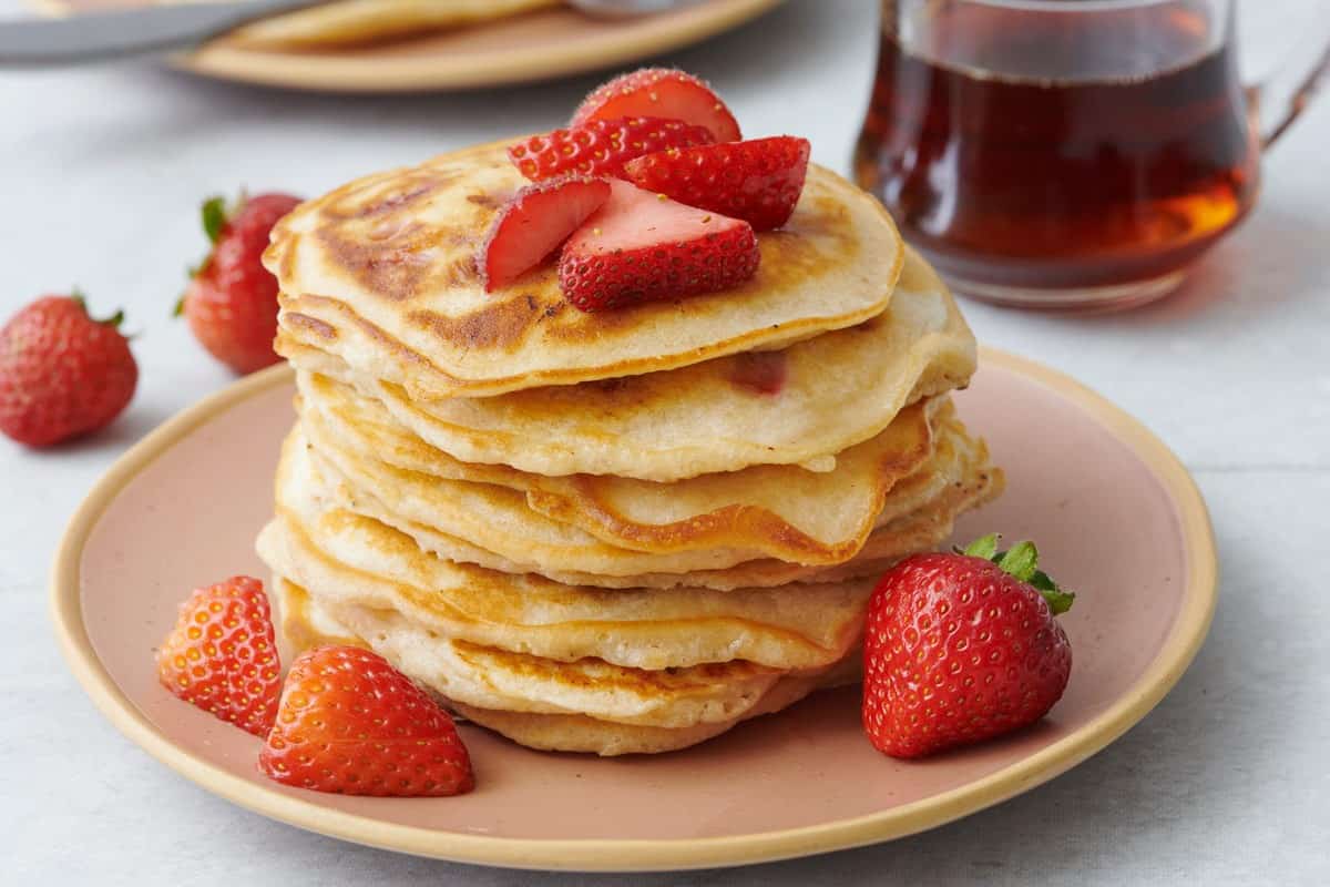 A stack of strawberry pancakes on a plate, topped with fresh strawberries with a container of syrup nearby.