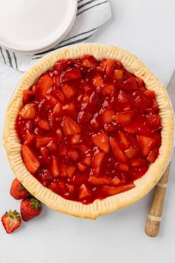 Strawberry pie in a pie tin with a spatula, fresh strawberries, small plates, and linen napkin nearby.