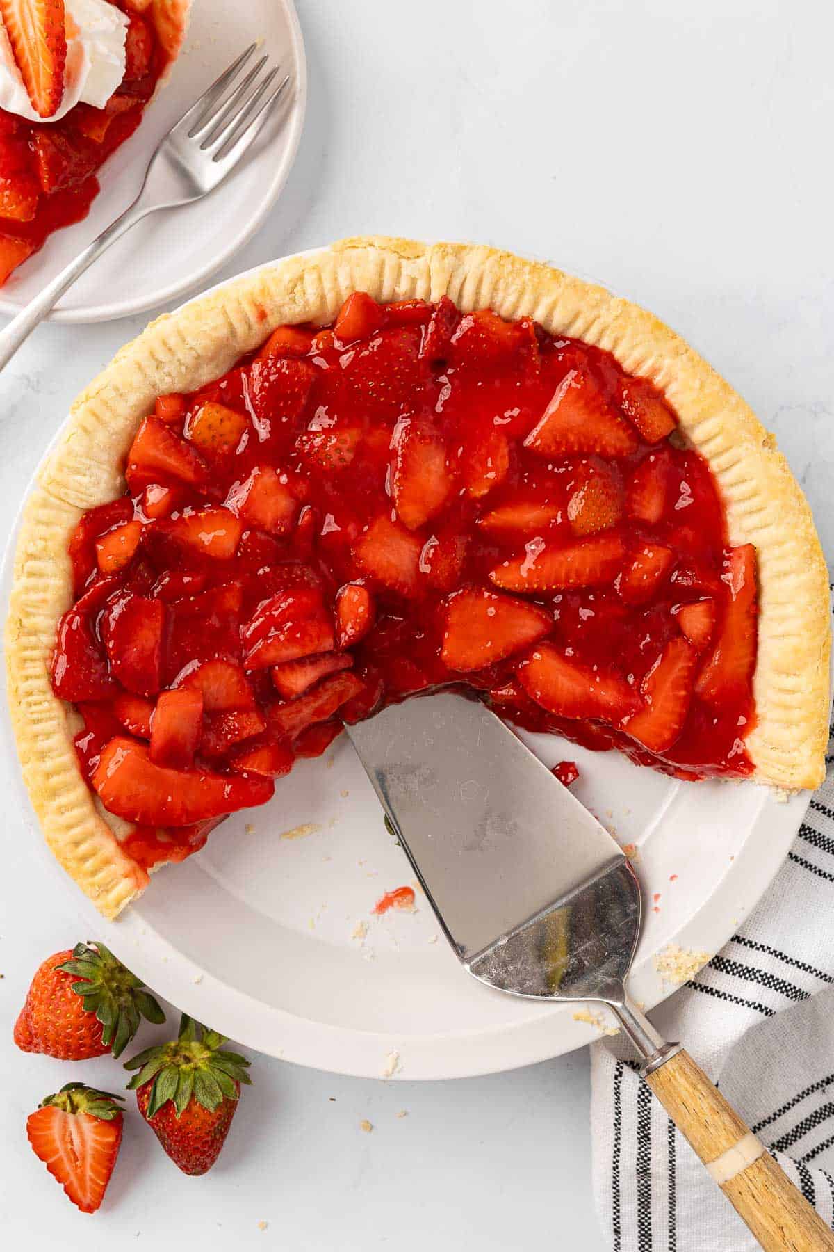 Strawberry pie in a pie tin with a few slices scooped out and a spatula ready to serve more; fresh strawberries and a slice on a small plate nearby.