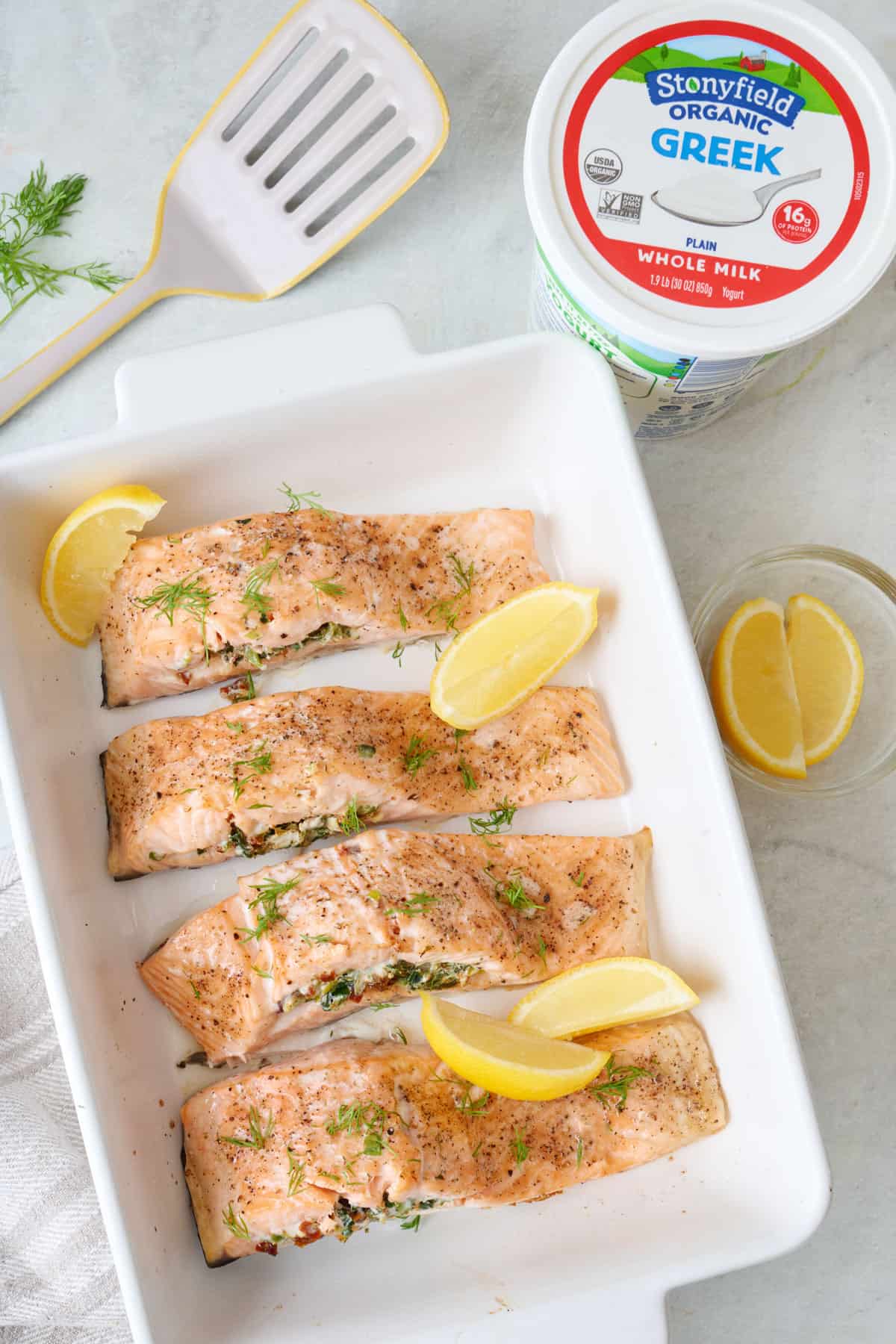 Baked stuffed salmon fillets in a white baking dish with lemon slices with a spatula and a carton of Stonyfield yogurt nearby.