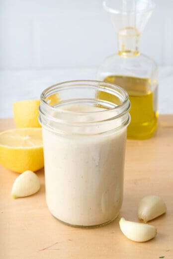 Homemade tahini dressing in a small jar with fresh lemon, garlic cloves, and oil around.