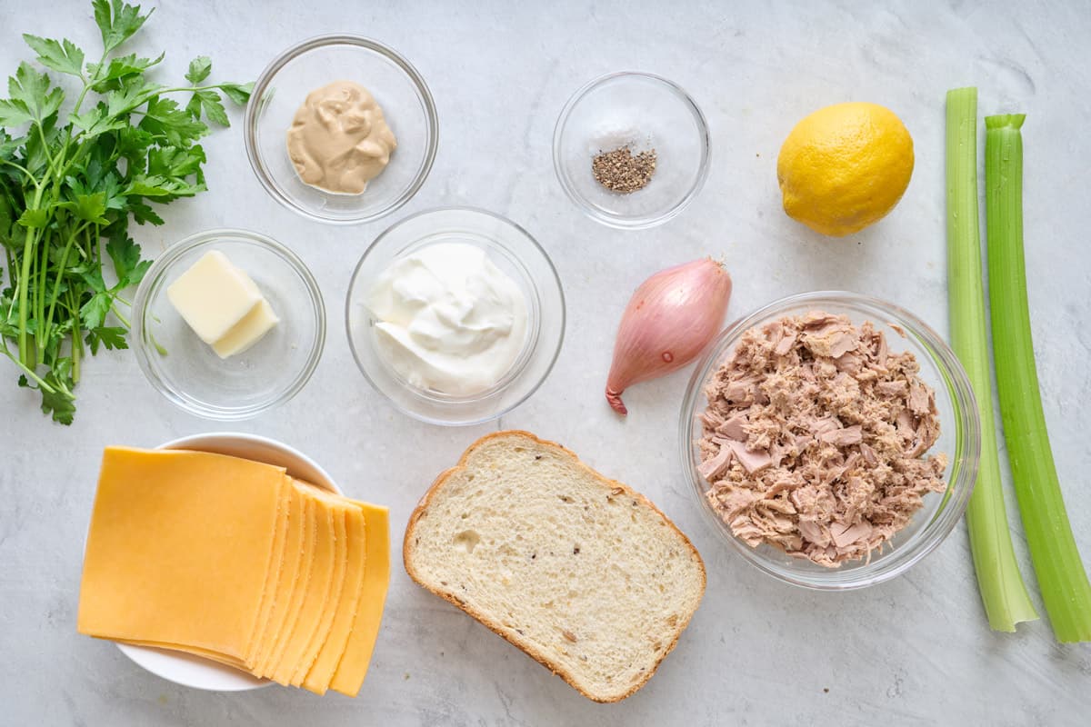 Ingredients for recipe in individual bowls: parsley, dijon mustard, butter, cheddar cheese slices, yogurt, bread slices, salt and pepper, shallot, lemon, tuna, and celery.