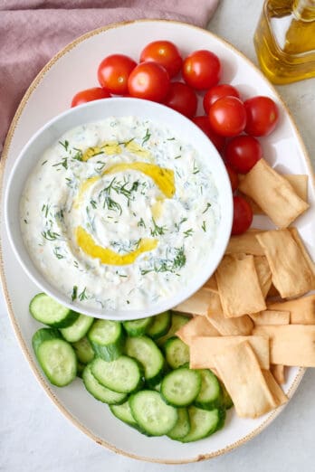 Tzatziki sauce on a platter surrounded by cherry tomatoes, sliced Persian cucumbers, and pita chips.