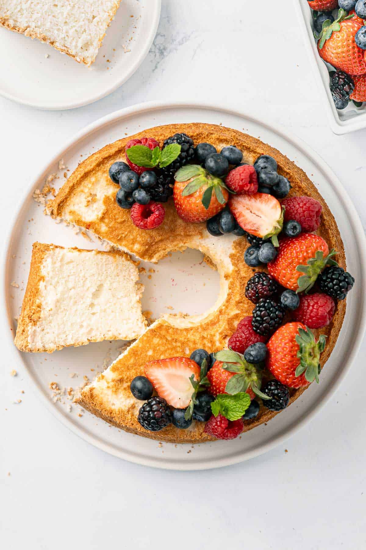 Angel food cake with a slice cut out and another slice on a plate nearby. Cake topped with fresh berries.