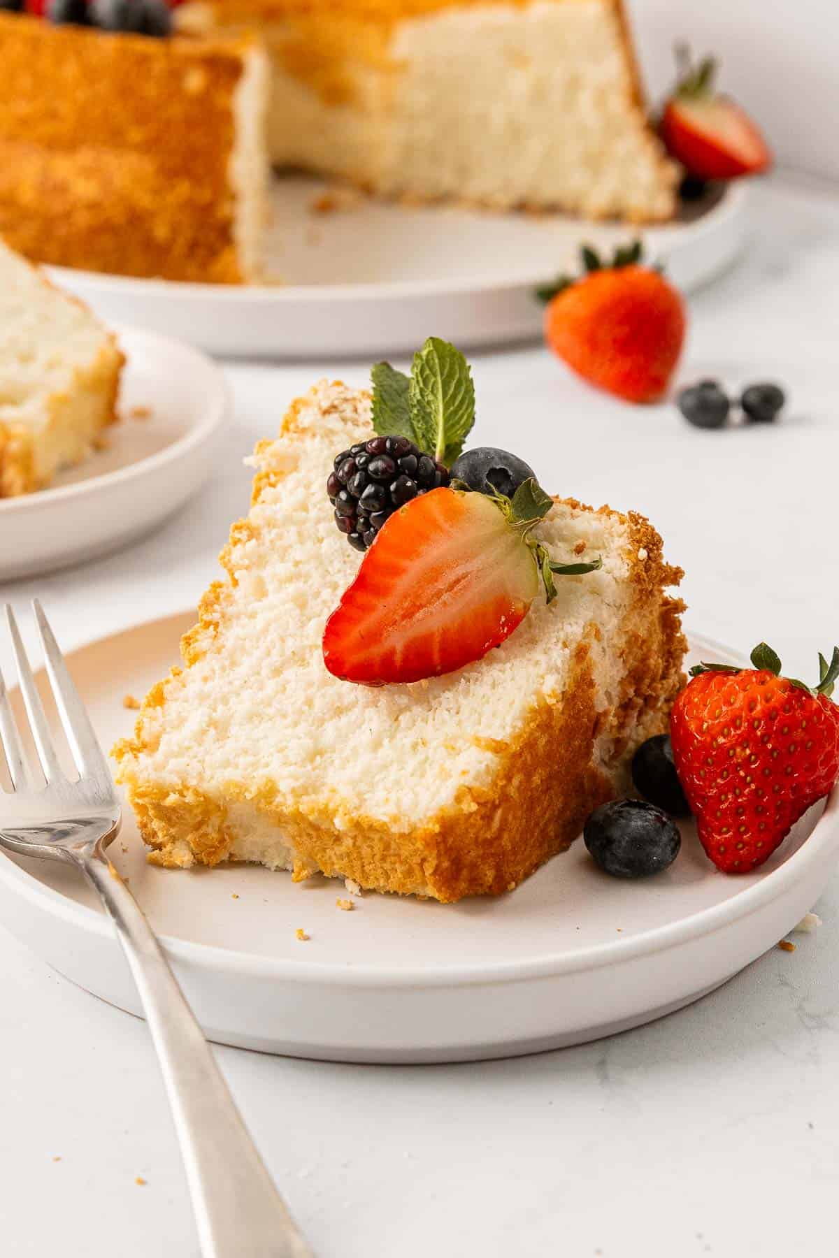 Slice of angel food cake on a plate topped with fresh berries and whole cake nearby.