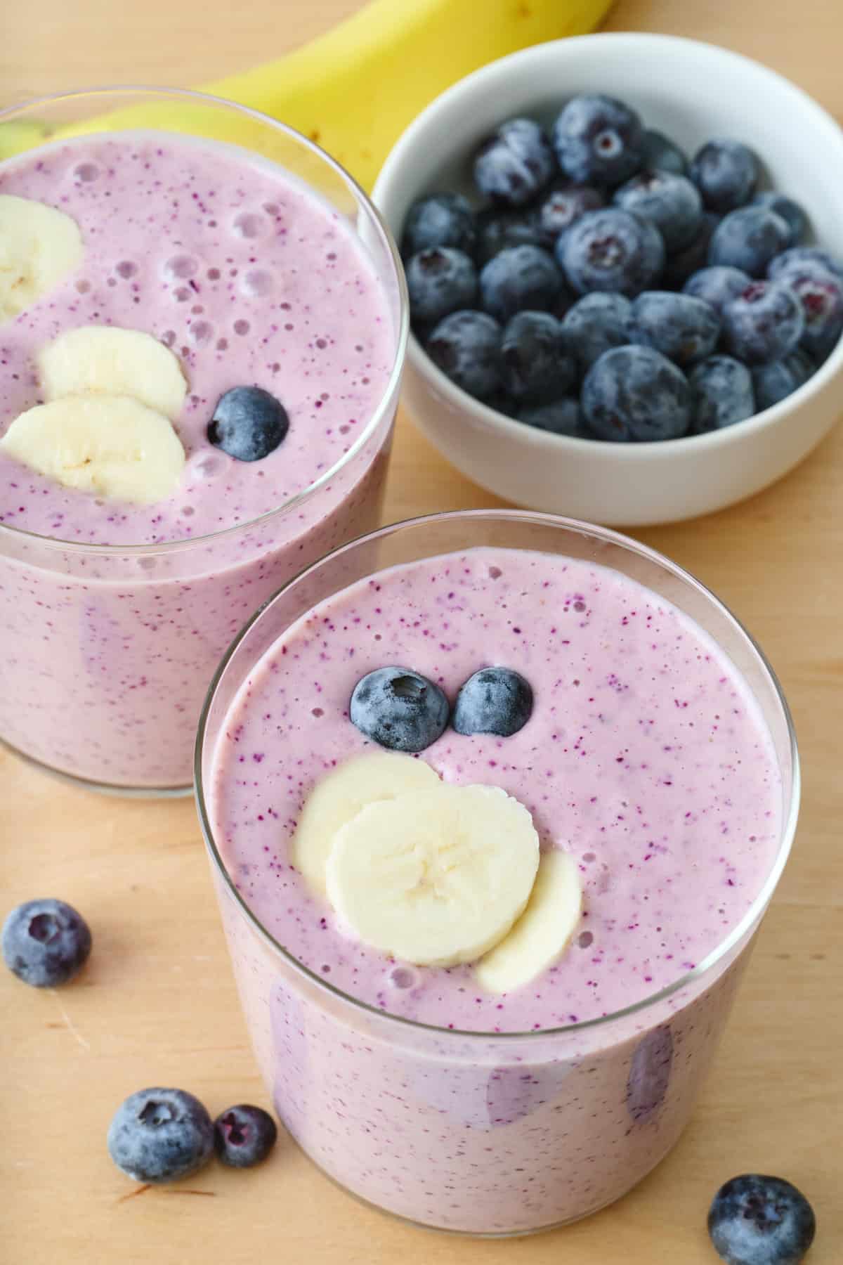 Two glasses of blueberry banana smoothie with a small bowl of fresh blueberries and a whole banana nearby.