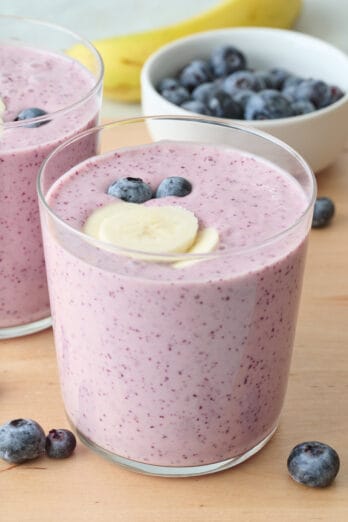 Small glass of blueberry banana smoothie garnished with fresh sliced bananas and blueberries with a bowl of fresh blueberries nearby.