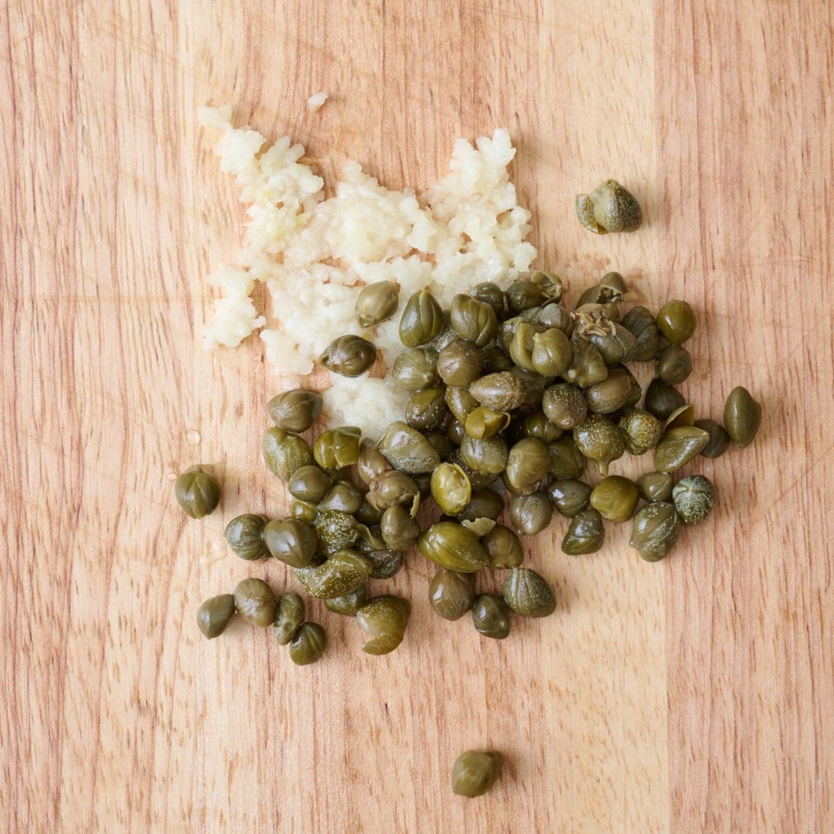 Minced garlic and capers on a wooden cutting board.