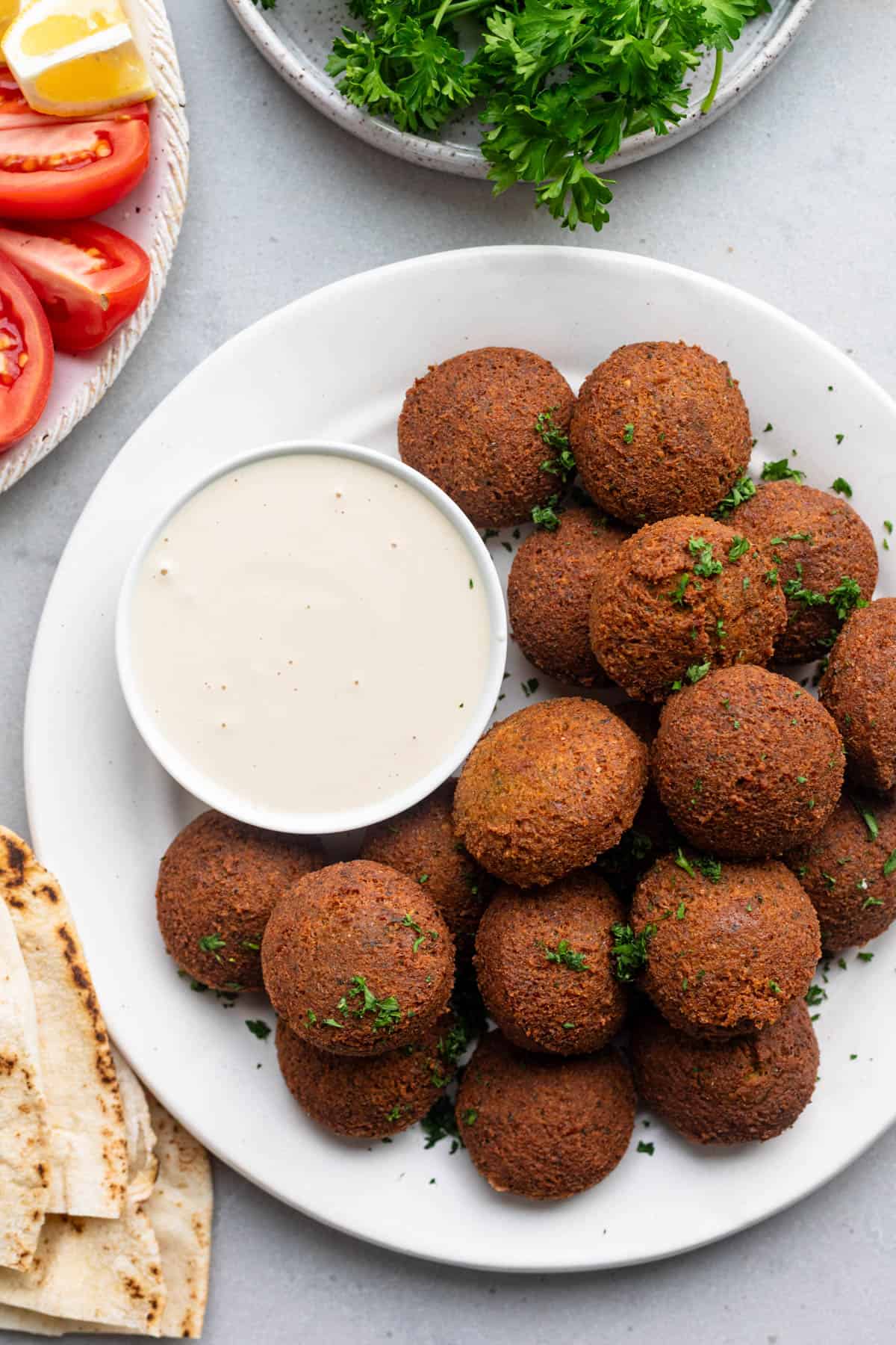 Crispy falafels piled on a plate with a small bowl of tahini sauce and fresh herbs, quartered tomatoes, and pita nearby.