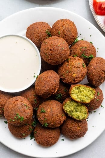 Plate of homemade falafel with one piece cut in half to show bright green bready insides. Small bowl of tahini sauce on plate for dipping.