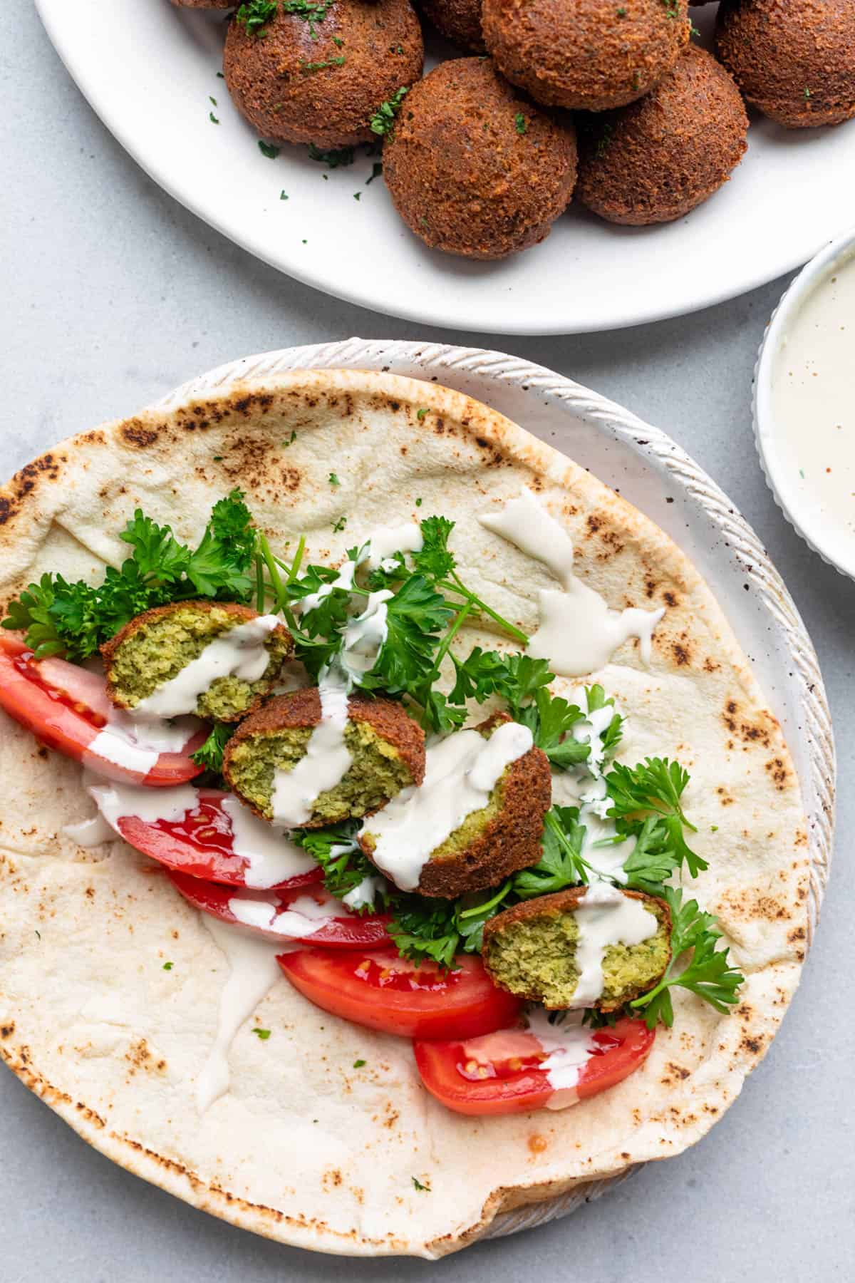 Two falafels torn in half and added to a pita with tomatoes, fresh parsley, and a drizzle of tahini sauce with plate of more falafel nearby.