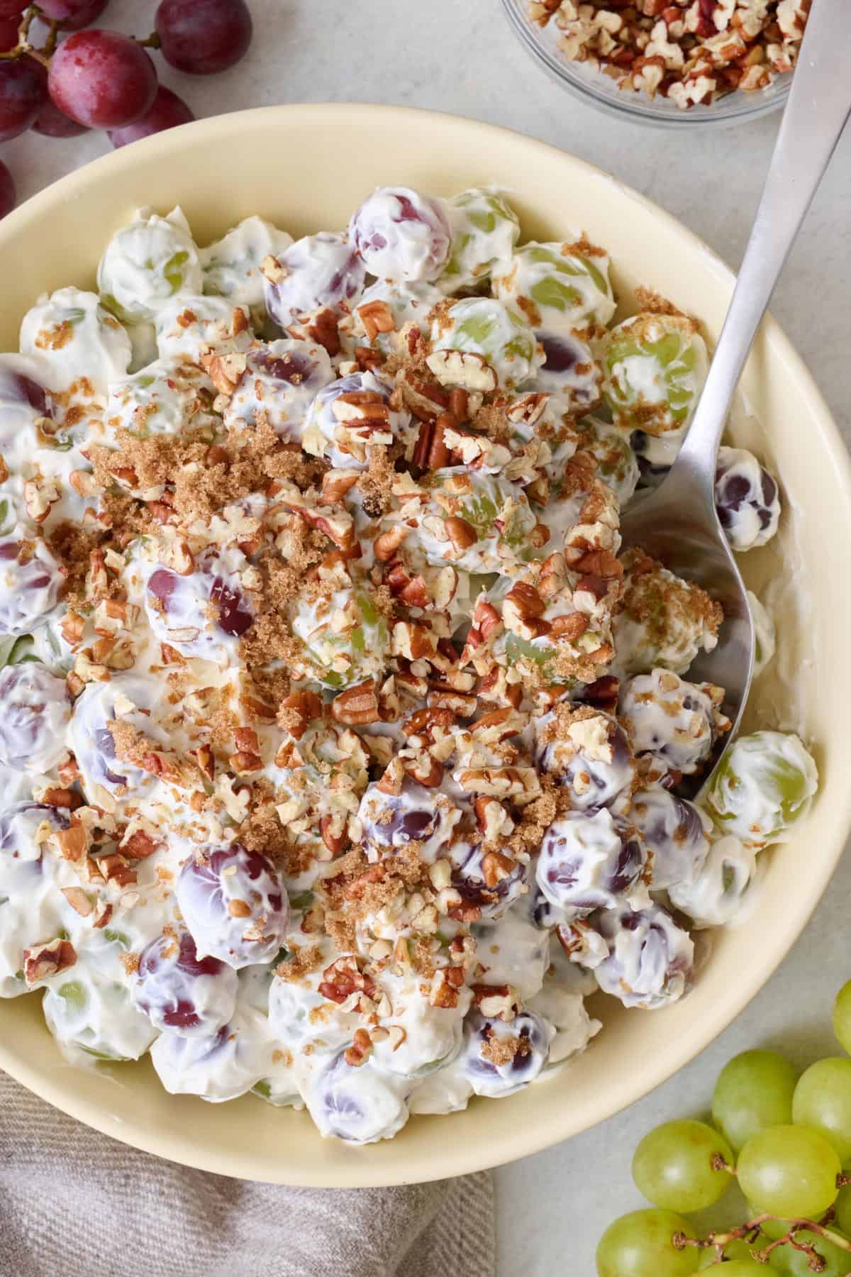 Bowl of grape salad topped with chopped pecans with spoon; fresh grapes and bowl of chopped pecans nearby.